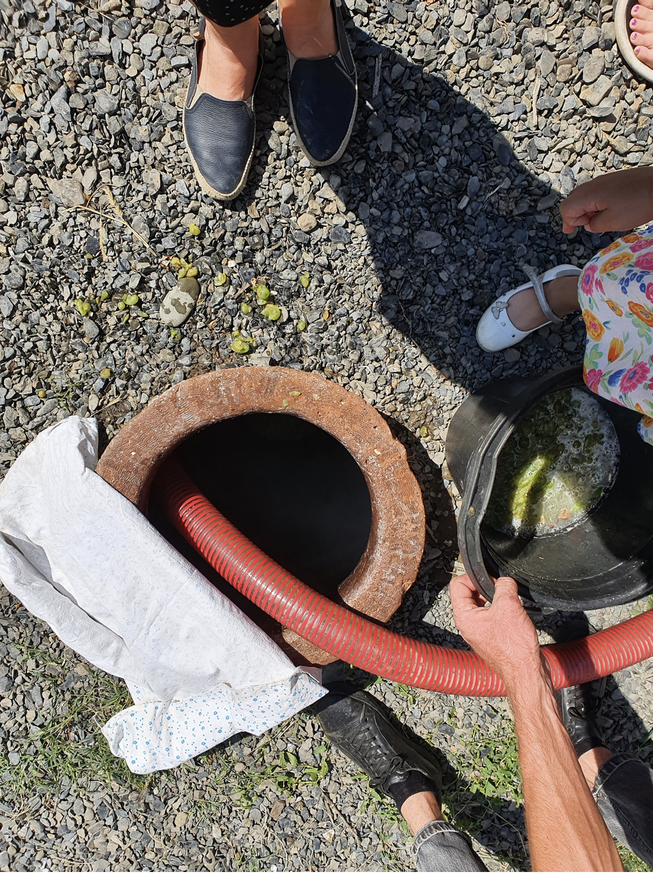 A hose directed into the mouth of a buried clay amphora, with the feet of onlookers visible