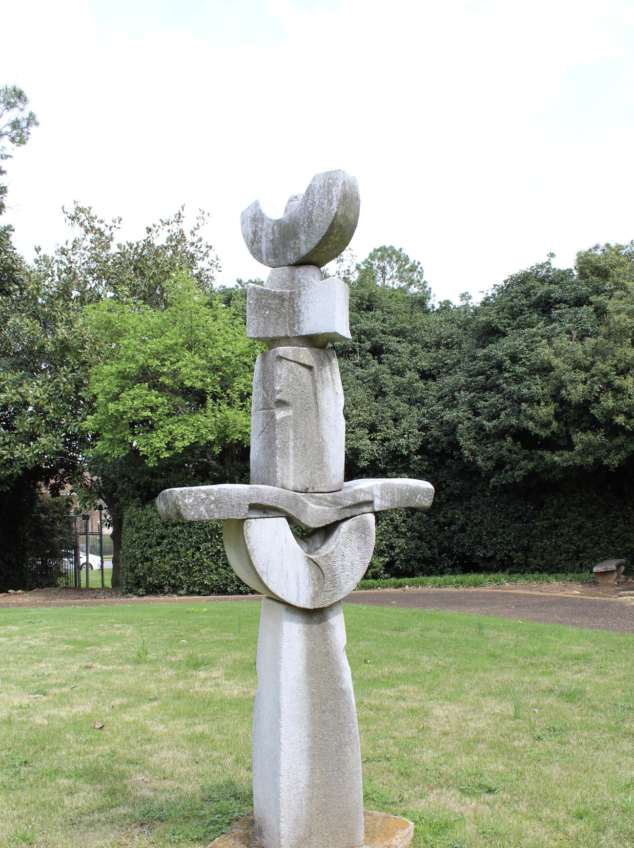 Photograph of an abstract granite sculpture standing in a turf lawn surrounded by tall shrubs